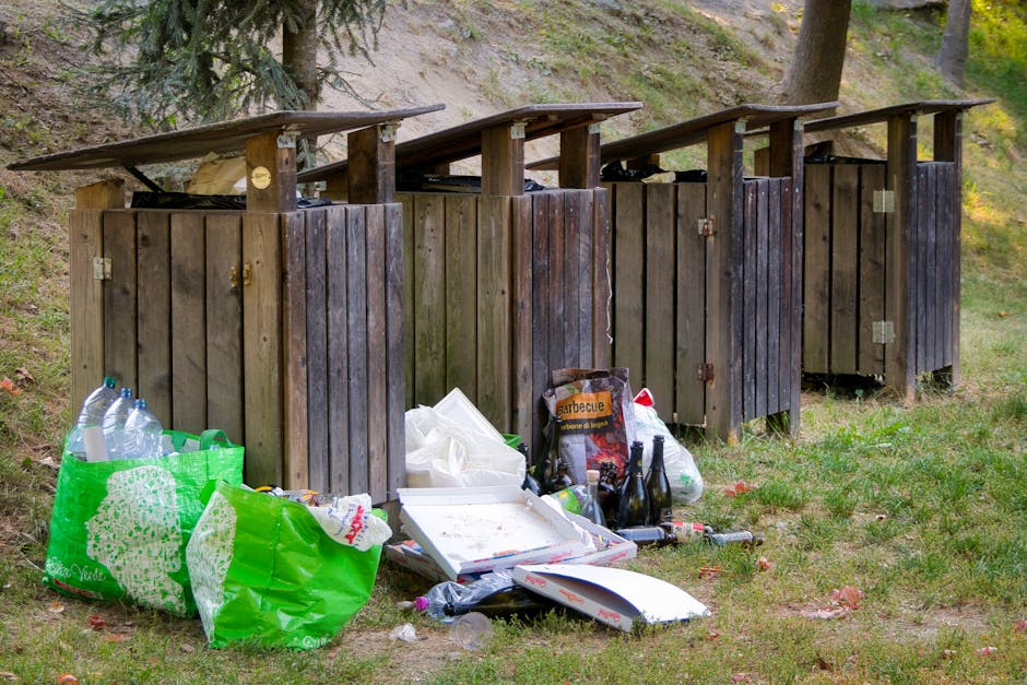 The image depicts a large landfill site with a steep, rubble-strewn slope filled with mixed waste materials, including plastic bags, cartons, and household rubbish. Several small yellow excavators are